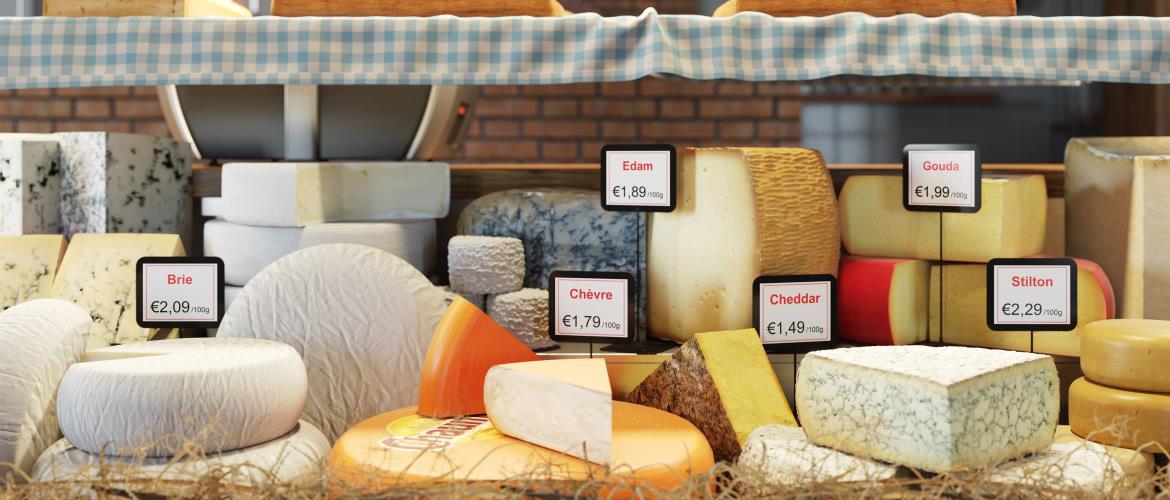 Une sélection de fromages (brie, chèvre, Edam, cheddar, Gouda et Stilton) est exposée dans une vitrine d'une boutique avec des étiquettes de prix en euros et des étiquettes de nom clairement identifiées pour chaque type de fromage.