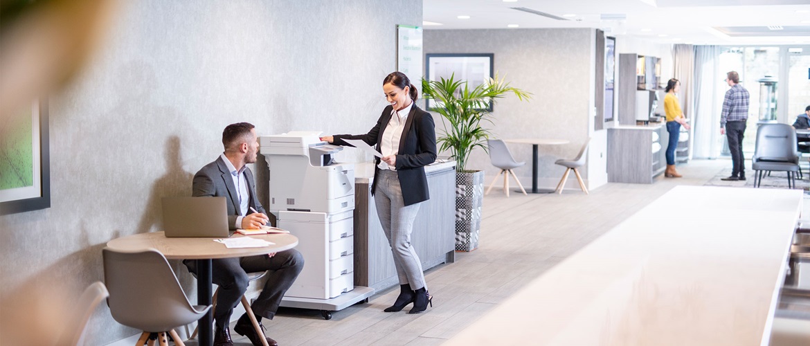 Une femme en costume se tenait devant une imprimante laser couleur Brother MFC-L9570CDWTT et un homme en costume est assis à la table d'à côté avec un ordinateur portable, des plantes, des gens au fond