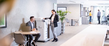 Une femme en costume se tenait devant une imprimante laser couleur Brother MFC-L9570CDWTT et un homme en costume est assis à la table d'à côté avec un ordinateur portable, des plantes, des gens au fond
