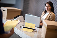 Femme aux cheveux bruns se tenant derrière la réception avec un ordinateur portable utilisant le scanner de documents portable DSmobile DS-740D de Brother avec des boîtes, un livreur